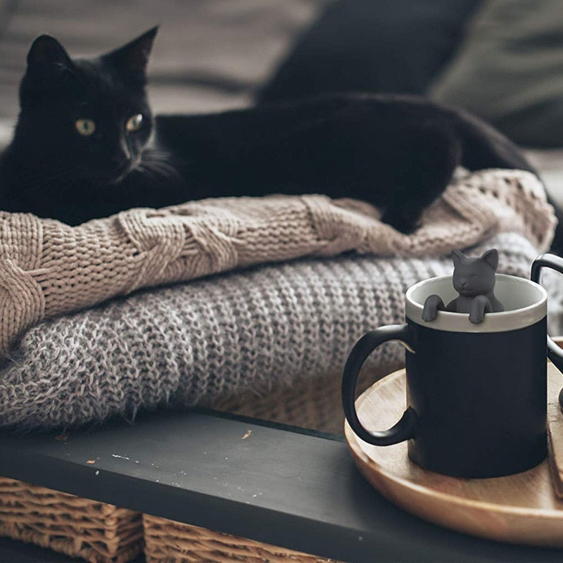 Cat-shaped infuser in a mug beside a resting black cat